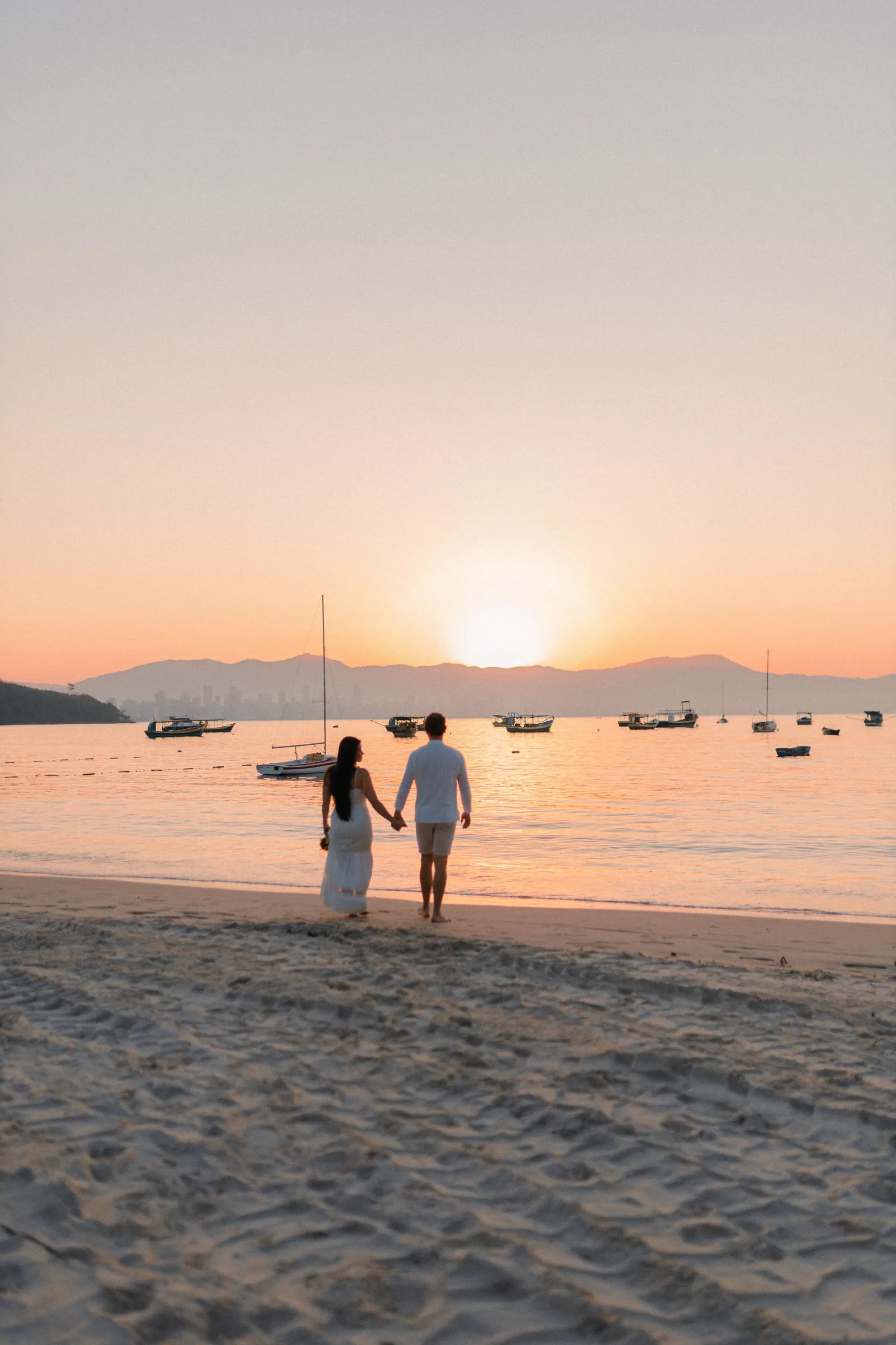 A couple in white walking hand in hand along the shore at sunset